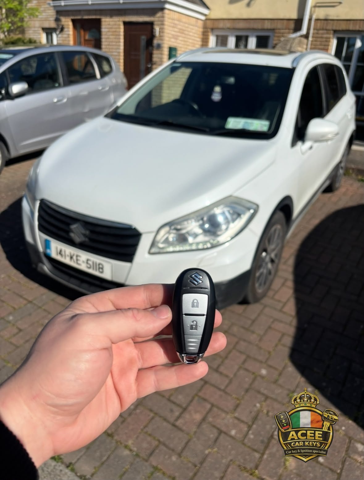Hand holding a black car key fob in front of a white Suzuki parked in a residential driveway