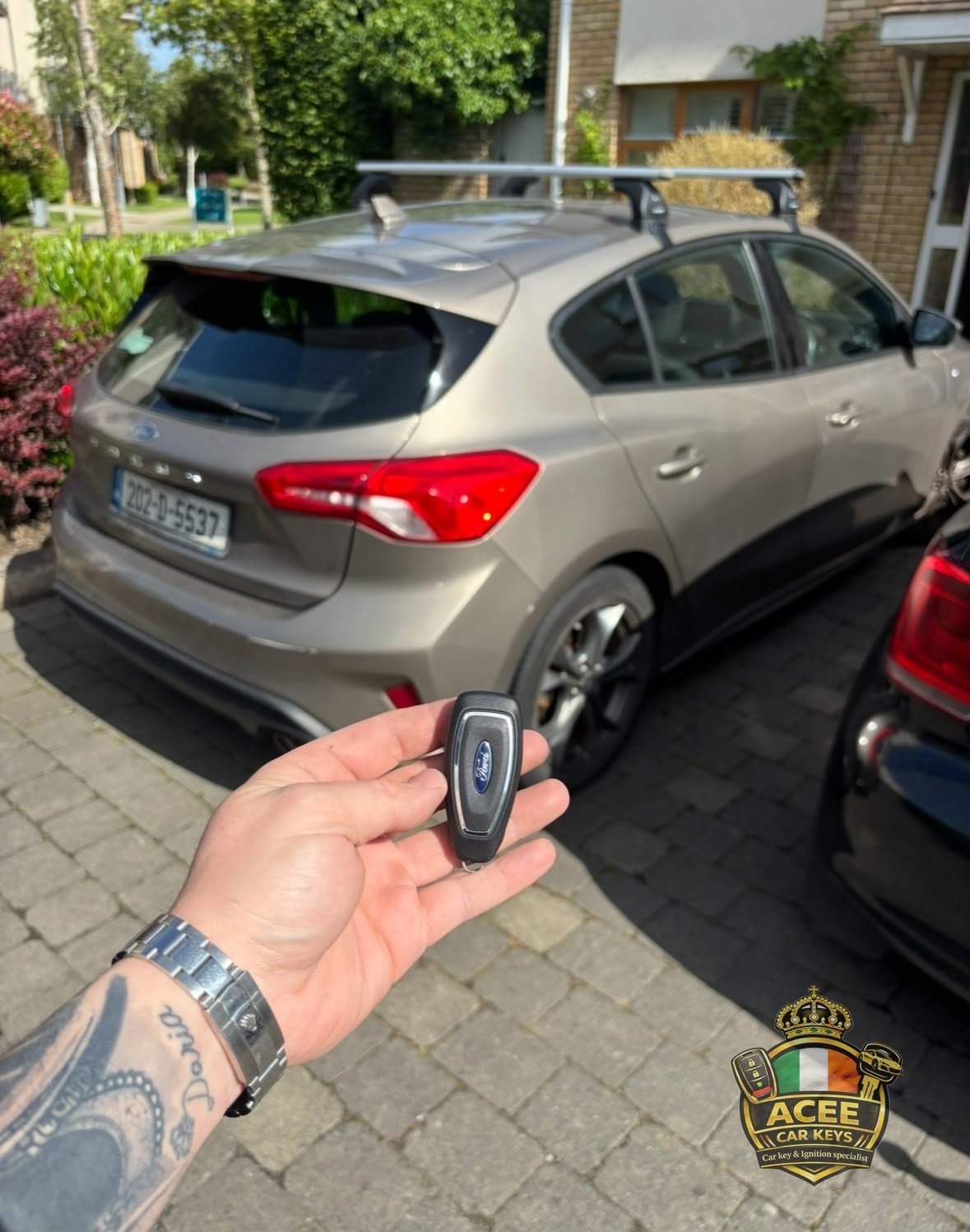 Hand holding a car key fob in front of a silver hatchback on a residential driveway