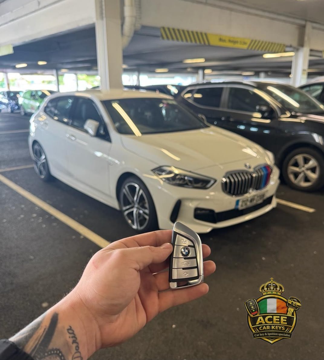Hand holding a BMW key fob in front of a white BMW in a multi-storey car park