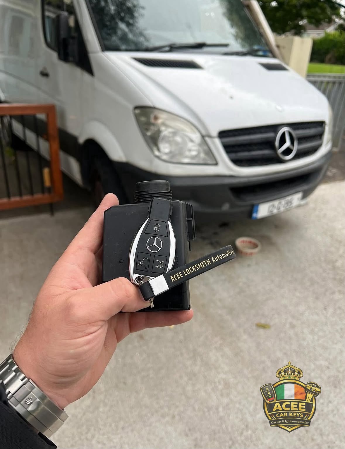 Hand holding a black Mercedes key fob with the Mercedes emblem in front of a white Mercedes van